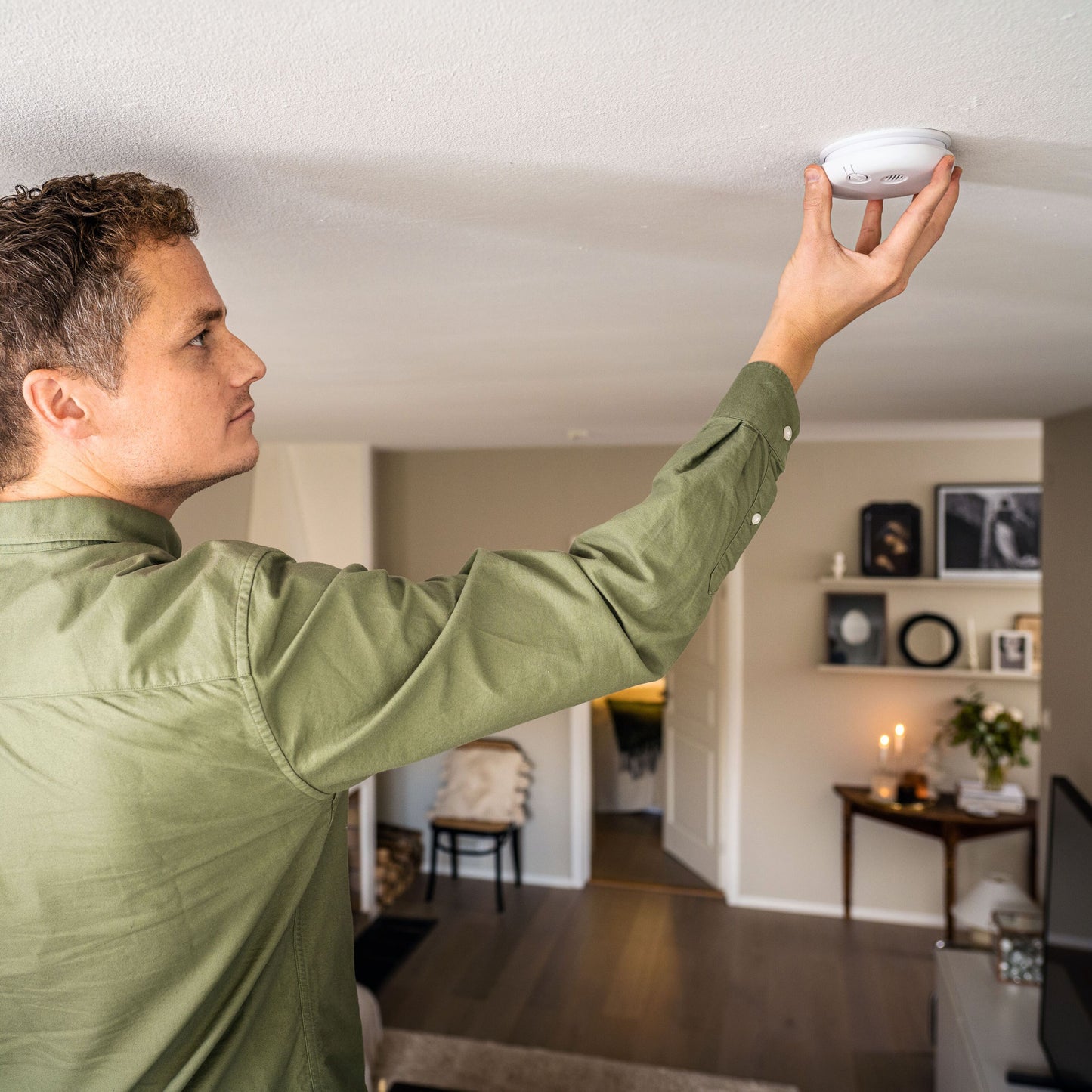 Housegard Pebble røgalarm monteres i loft af stue, grøn trøje, hylder, billeder og lysestage synlige i baggrunden.