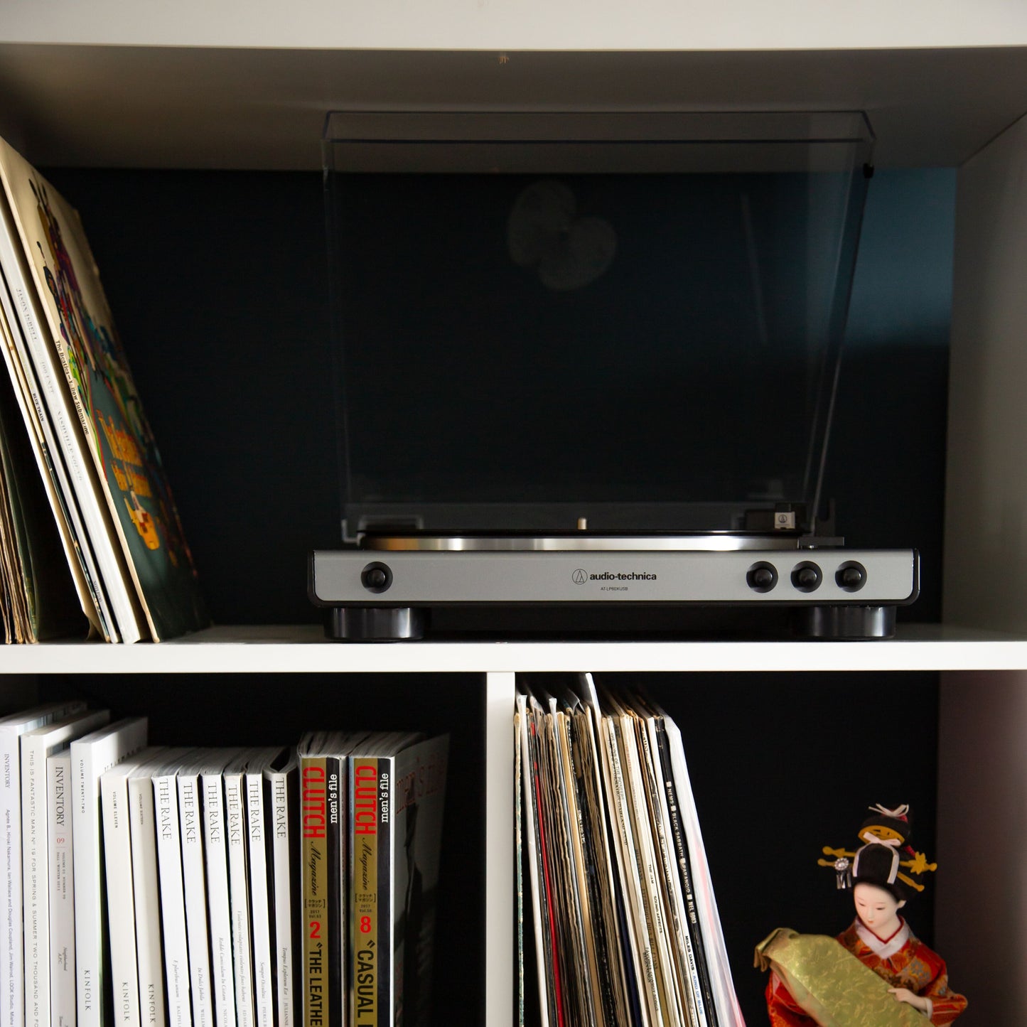 Audio Technica AT-LP60XUSB vinylafspiller med lukket låg på hylde over LP-plader og magasiner, dukke ses på nederste hylde.