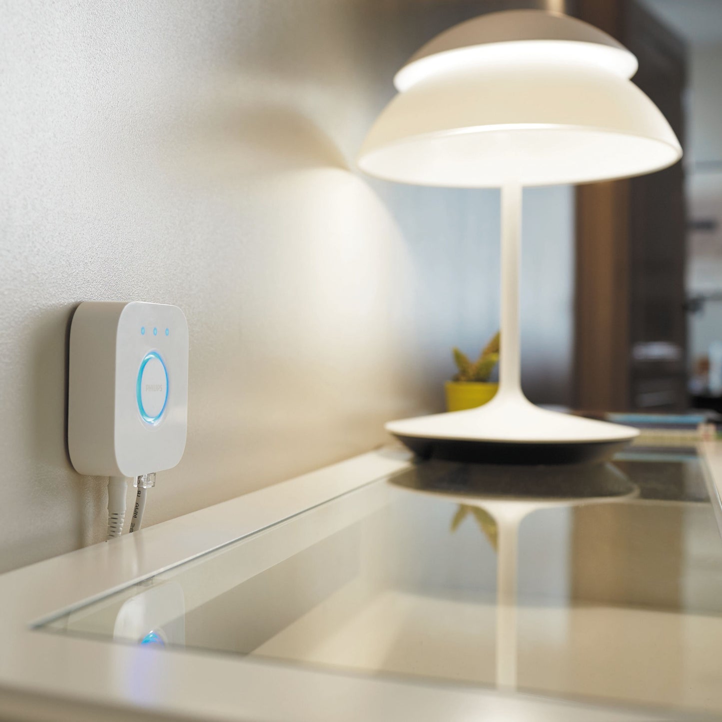 Philips Hue Bridge kompakt, vægmonteret ved hvid bordlampe og plante på glasbord, synlig forside og detaljer.