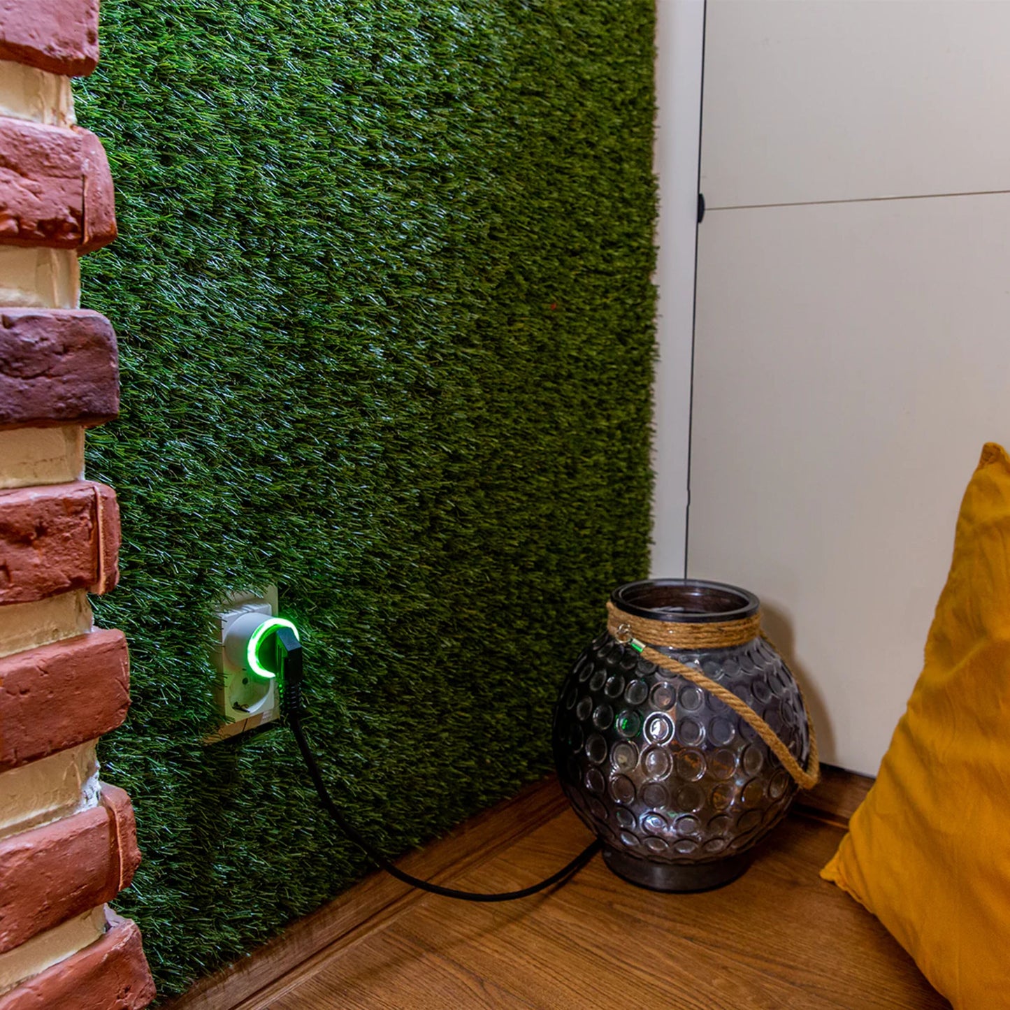 Shelly Plug S Gen3 smart stik i væg med kunstgræs, synlig glaskrukke og gul pude på trægulv, delvis murstensvæg.