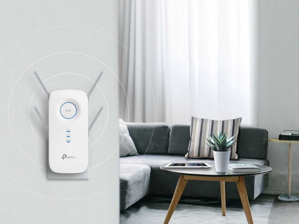 TP-Link AC2600-repeater med MU-MIMO, vægmonteret nær grå sofa og rundt træbord, hvidt design.