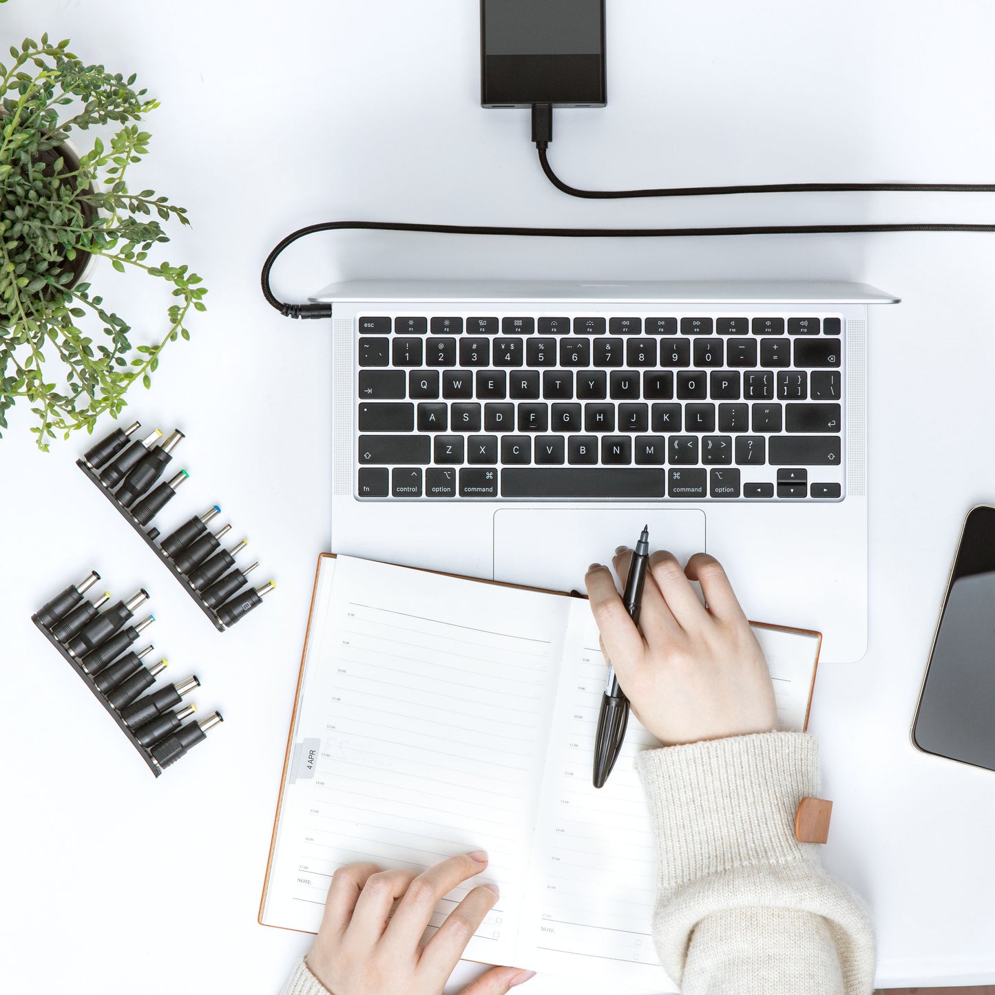 Plexgear Universal USB-C til DC-kabel 100 W tilsluttet laptop ved siden af telefon, oplader og strømadaptere på skrivebord.