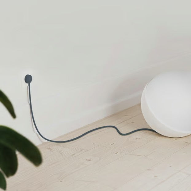 Rund hvid lampe med lang sort ledning tilsluttet NORM K stikprop blå med jord på lyst trægulv nær væg, grøn plante ses.