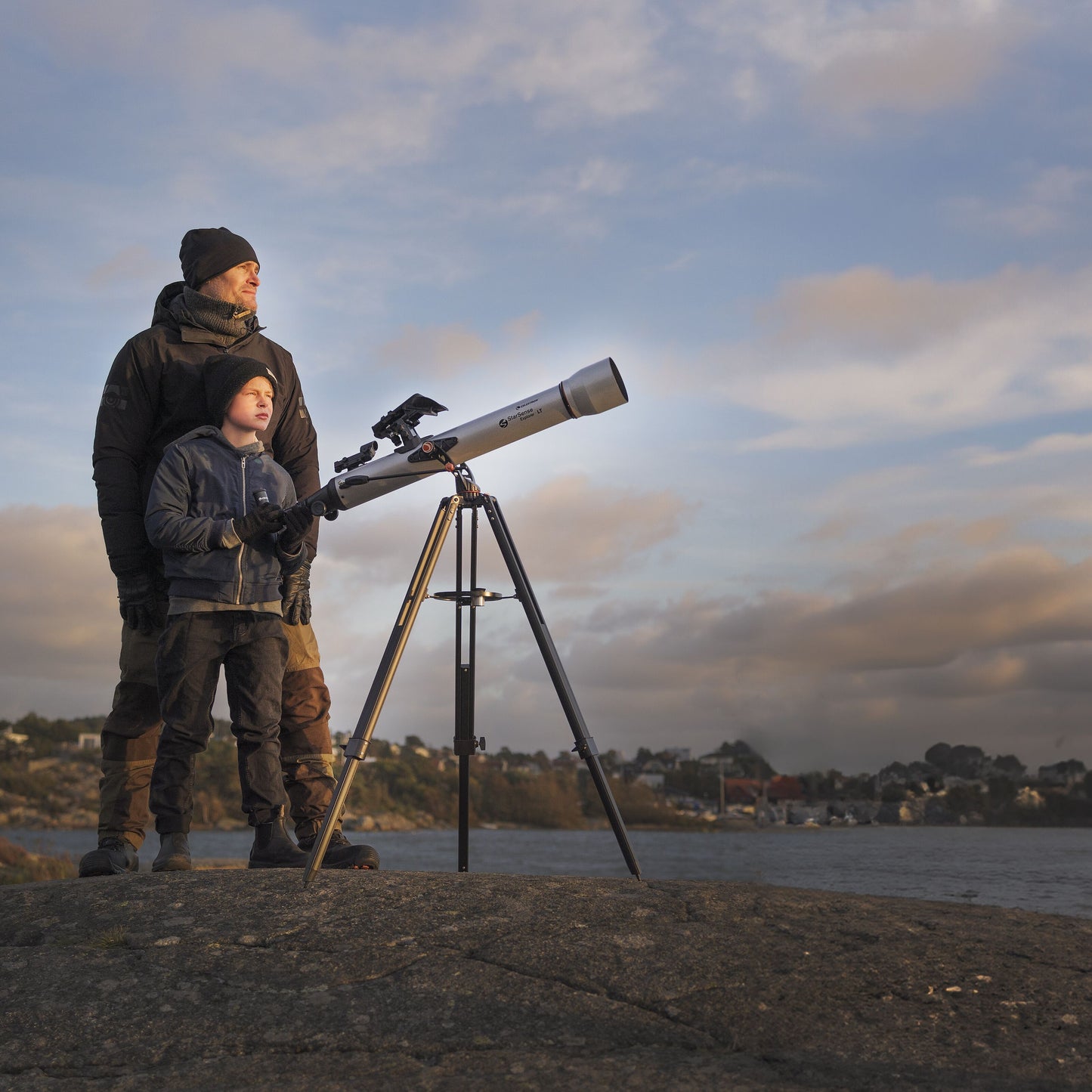 Celestron Powerseeker 60 AZ teleskop på azimutal montering holdt af barn ved klippekyst, huse og vand i baggrunden.