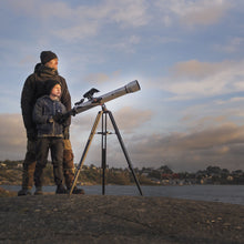 Celestron Powerseeker 60 AZ teleskop på azimutal montering holdt af barn ved klippekyst, huse og vand i baggrunden.