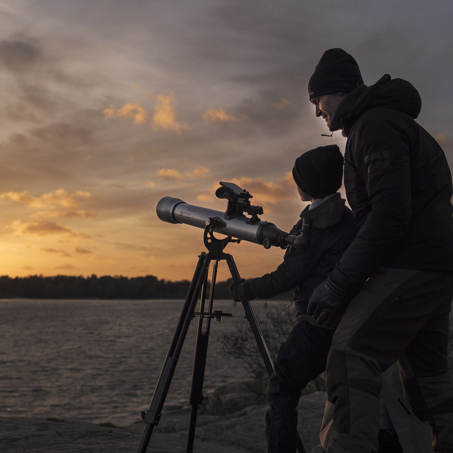 Celestron Powerseeker 60 AZ teleskop på azimutal montering med to personer i overtøj ved vand og træer i baggrunden.