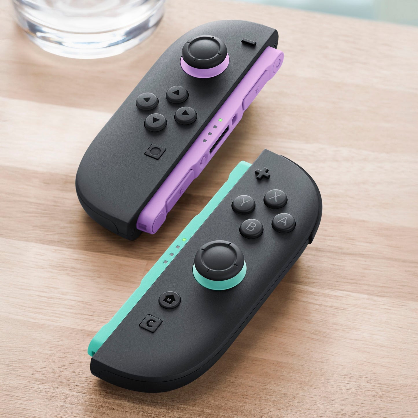 Nintendo Joy Con 2 Par controllere i lys lilla og lys grøn på træbord, delvist synlig vandglas i baggrunden.