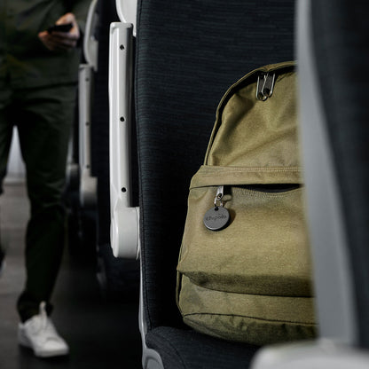 Khaki rygsæk med sort Chipolo One Spot Bluetooth nøglefinder (Apple Find My) monteret, på mørkt bus- eller togsæde.