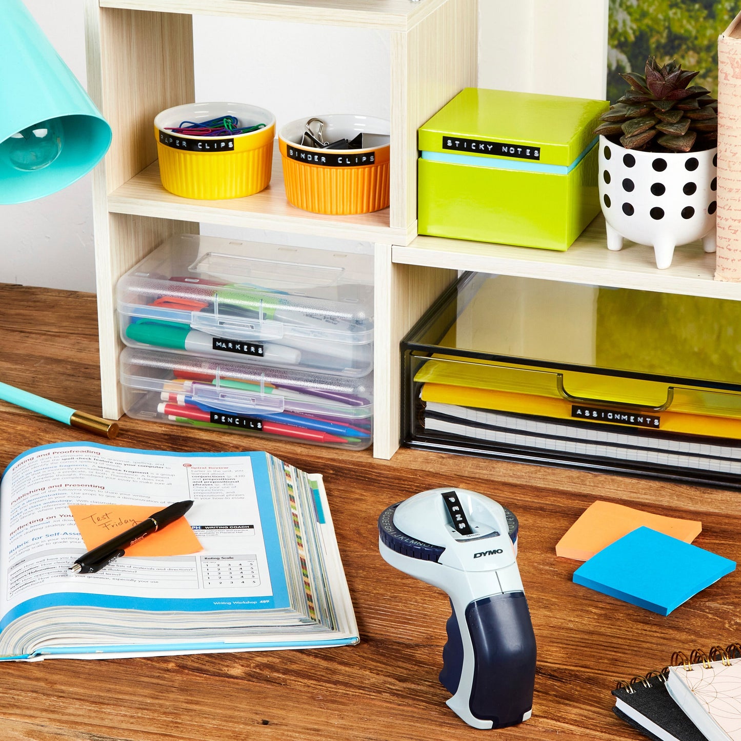 Dymo Omega Prægemaskine, farvede kontorartikler, mærkede bøger, sticky notes, kuglepenne, åben notesbog.