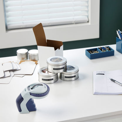 Dymo Omega Prægemaskine, 4 mærkede metaldåser, papkasse, kuverter, glas, penne og blå clipsbeholder.