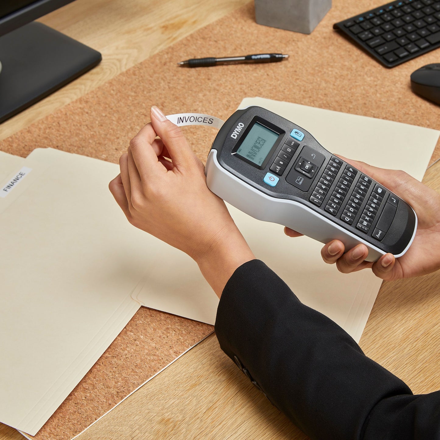 Dymo Labelmaskine 160 med QWERTY-tastatur printer "INVOICES"-etiket, filmappe i hånden, skrivebordsmiljø.