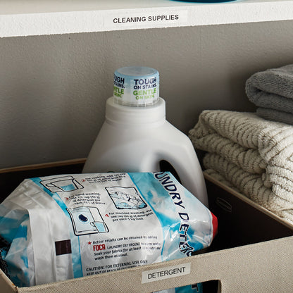 Hylde med skilt CLEANING SUPPLIES, kurv med etiketten DETERGENT, vaskemiddel, 3-pak vaskepods og foldede håndklæder.