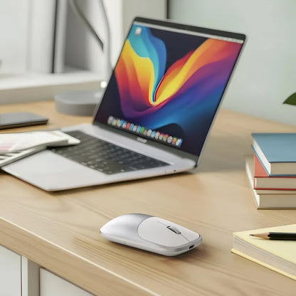 Satechi Slim EX Mus, sølvfarvet ved siden af en sølvfarvet bærbar computer med farverig skærm og USB-C på et skrivebord af træ.