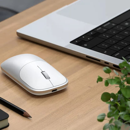 SATECHI Slim EX mus i sølv med Bluetooth 5.3 ved siden af en bærbar computer, en sort notesbog, en blyant og en lille grøn plante.
