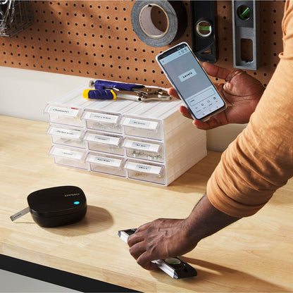 Dymo Letratag 200B trådløs etiketprinter bruges med smartphone ved værksted, skuffer og værktøj synlige i baggrunden.
