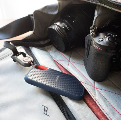 Sandisk Portable SSD Extreme 4TB blå/rød fastgjort på sort karabinhage ved kamera og objektiv i åben grå kamerataske.