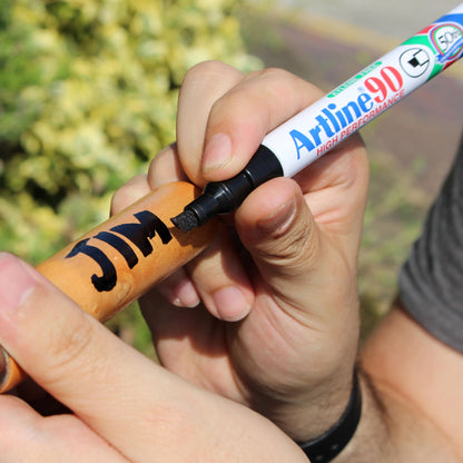 Artline Tuschpennor 90 Sort marker 4-pack skriver "JIM" på lysebrun overflade, uskarp grøn baggrund.