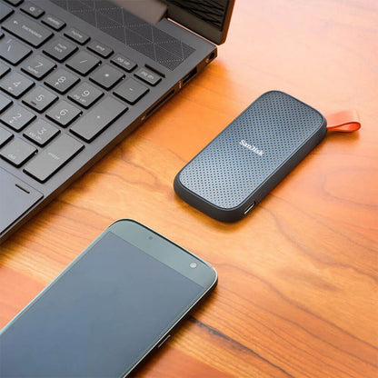 Sandisk bærbar SSD-harddisk 2TB med perforeret top og orange løkke mellem laptop-tastatur og smartphone.