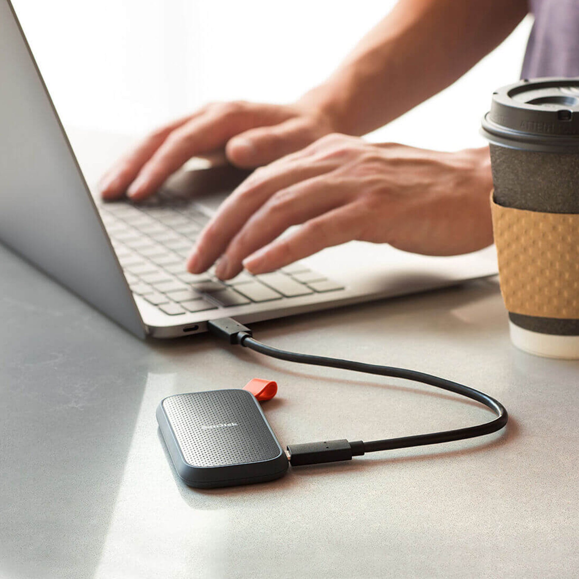 Sandisk Bærbar SSD-harddisk 2 TB tilsluttet laptop via USB, takeaway kaffekop ved siden af på bord.