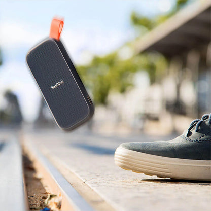 SanDisk bærbar SSD 2TB med orange løkke, vist tæt på en sneaker udendørs.