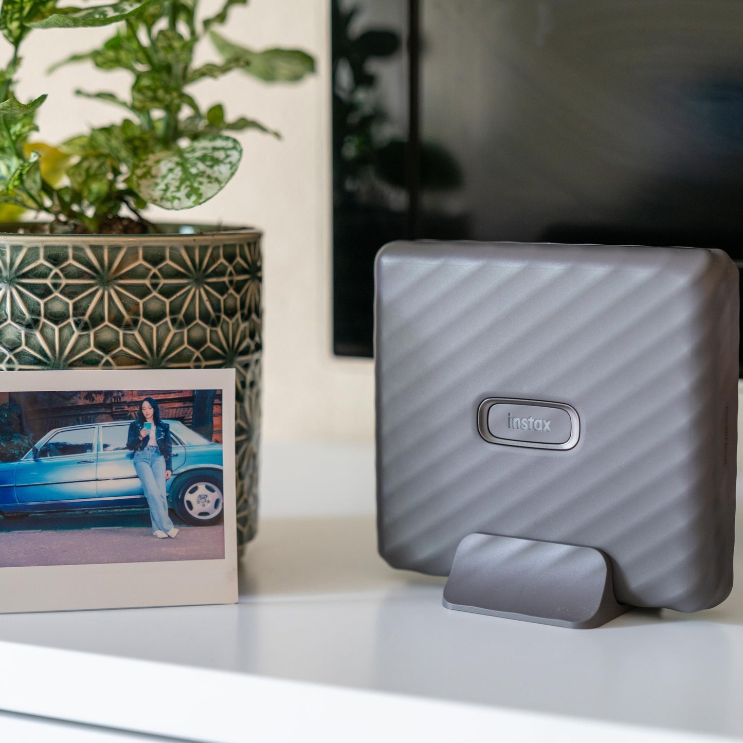 Fujifilm Instax Link Wide smartphone printer hvid på stander, instant foto og grøn plantepotte synlige.