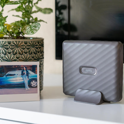 Fujifilm Instax Link Wide smartphone printer hvid på stander, instant foto og grøn plantepotte synlige.