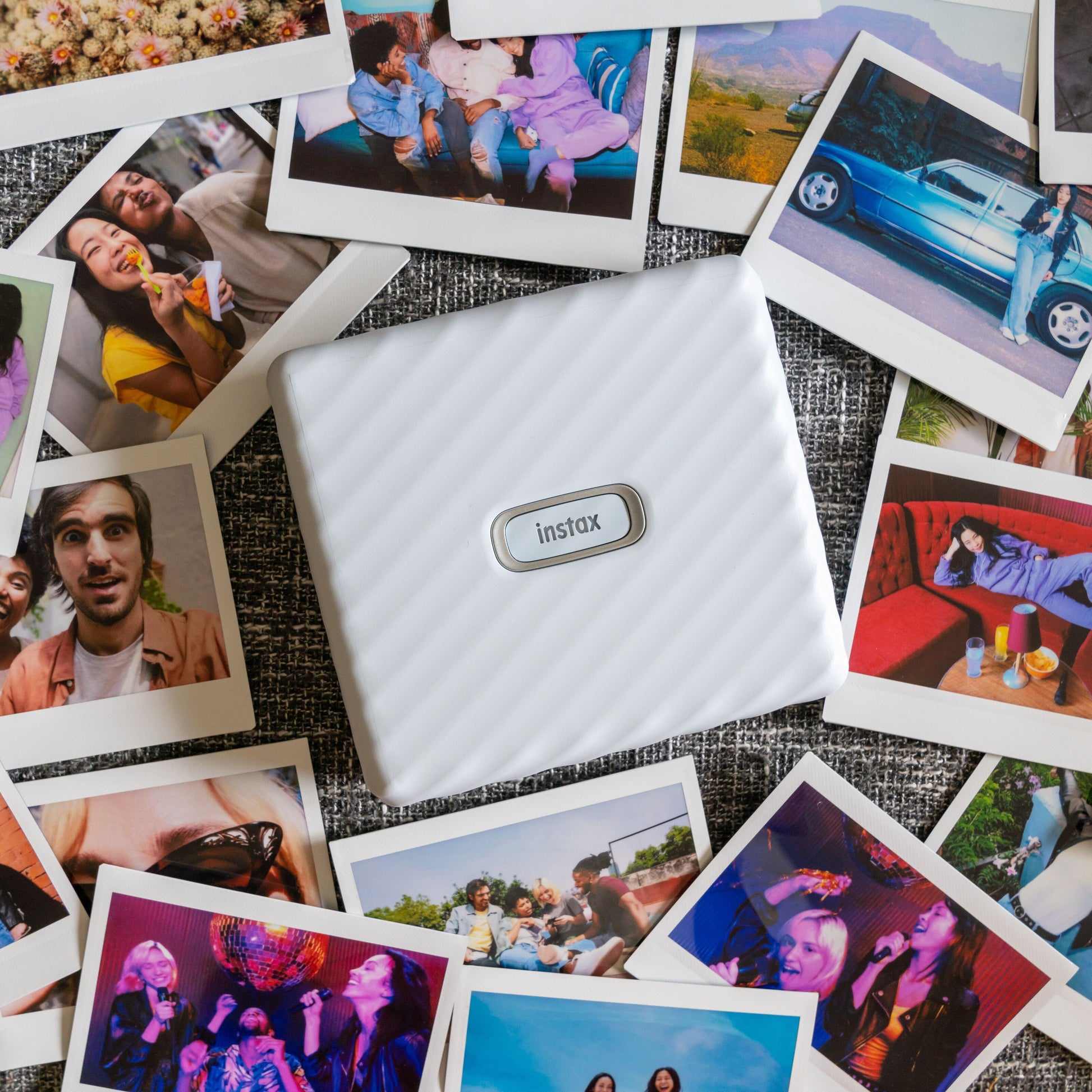 Fujifilm Instax Link Wide Hvid printer omgivet af farverige Instax Wide-fotos af personer, landskaber og begivenheder.