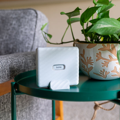 Fujifilm Instax Link Wide hvid smartphone printer på grøn bordplade ved siden af potteplante og grå polstret stol.