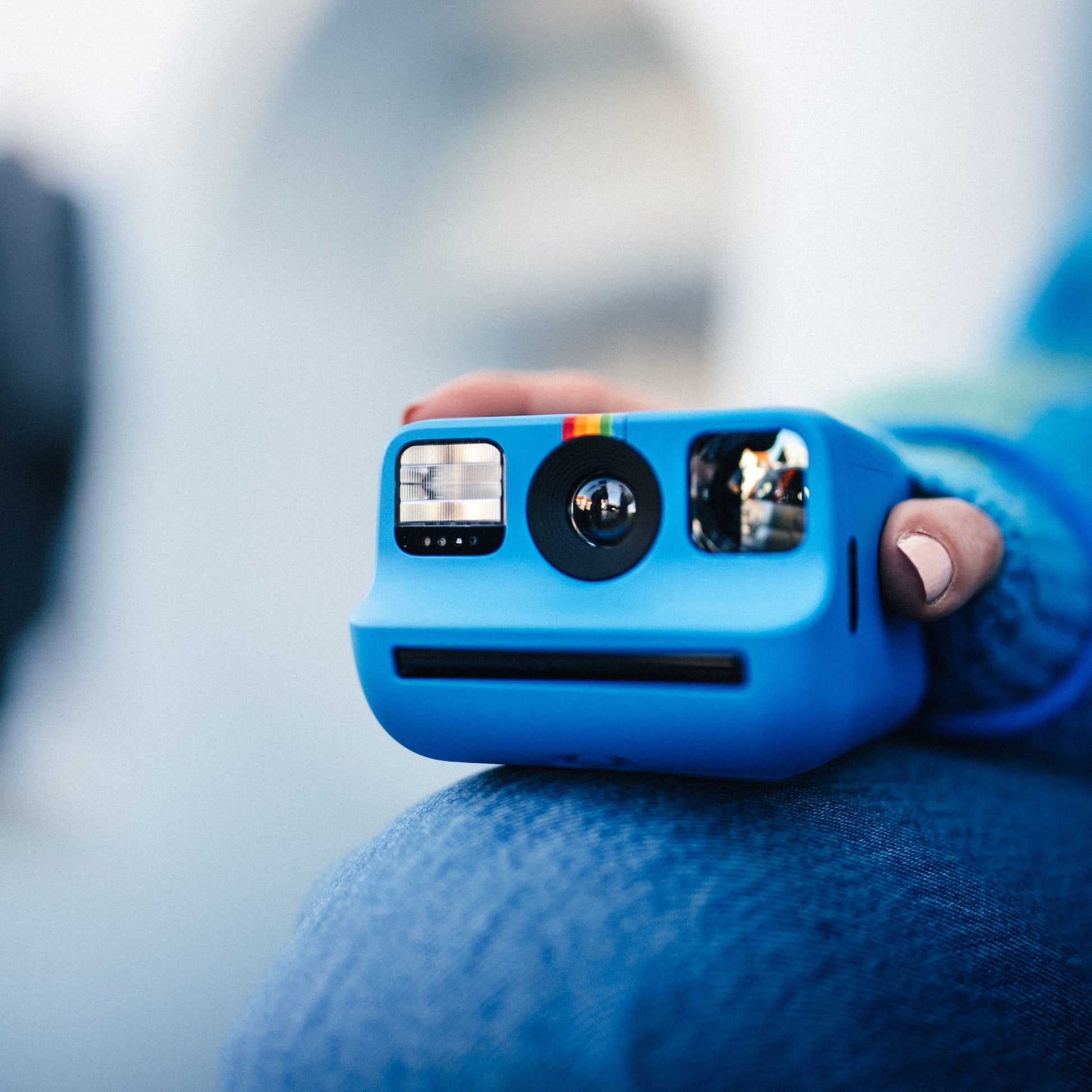 Polaroid Go Gen2 Instant kamera i hvid holdes over et knæ med denim, skarpt fokus på kameraets front og linse.