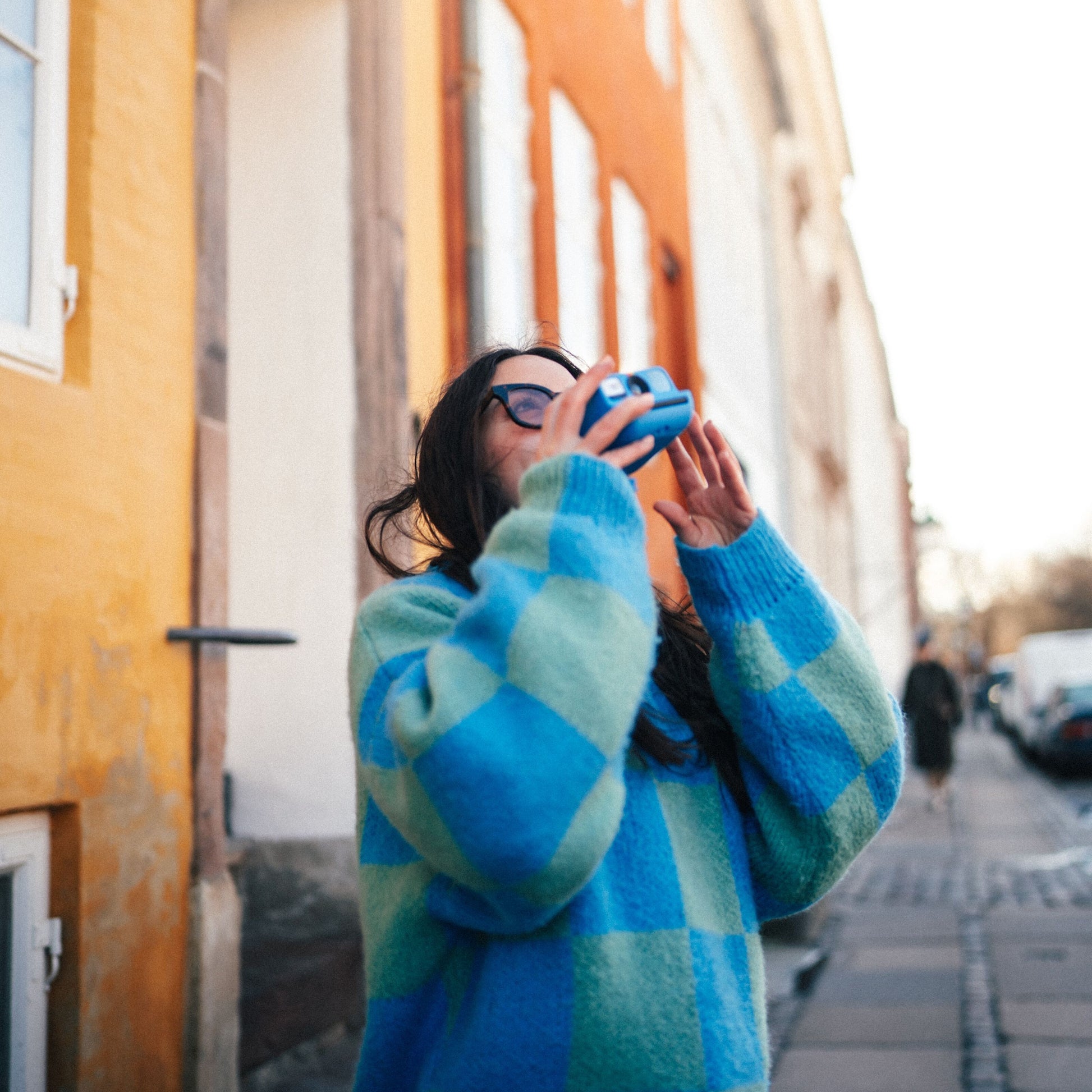 Polaroid Go Gen2 Instant kamera i brug, blå/grønternet sweater, solbriller, bybaggrund med orange/hvide bygninger.