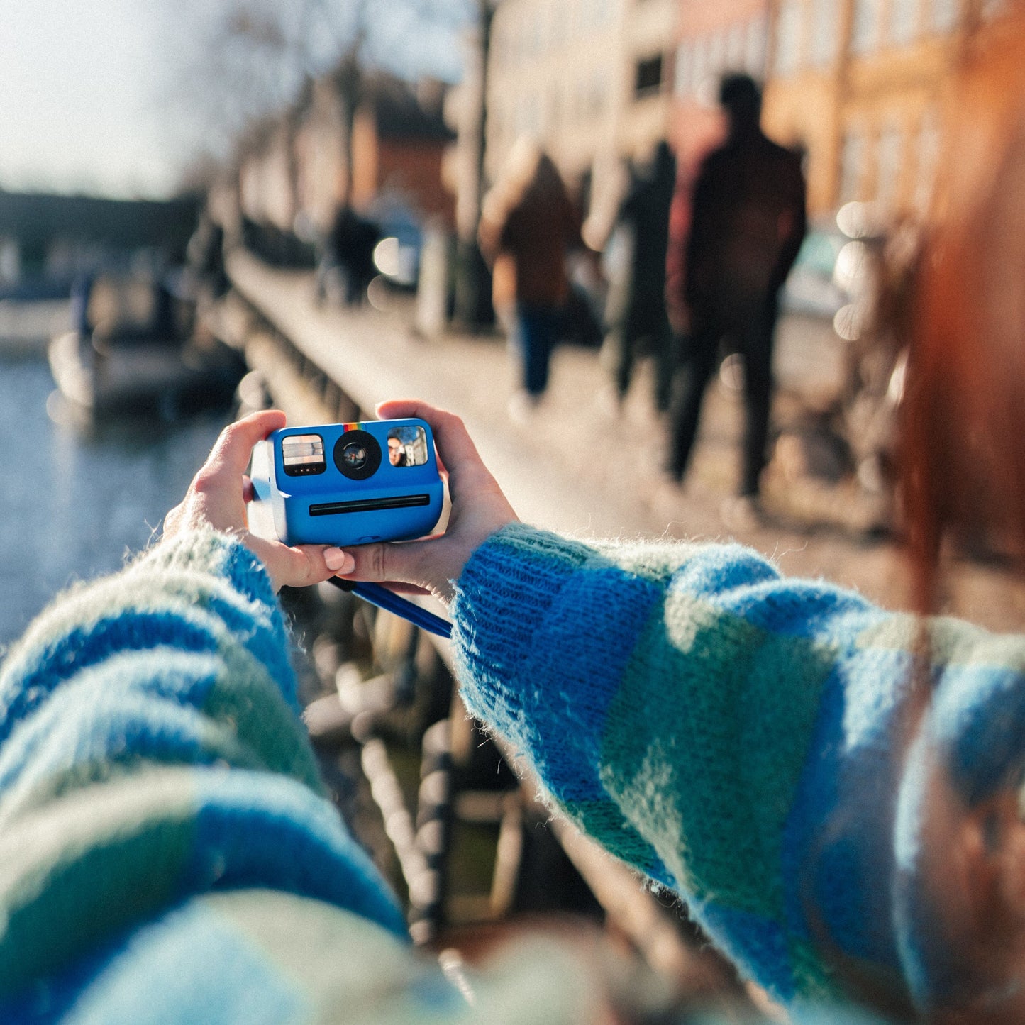 Polaroid Go Gen2 Instant kamera i brug, blå/grøn-stribet trøje, snapshot af personer ved vandet i sollys.