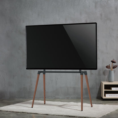 TV gulvstand Bauhaus-design til 60-75" fladskærm, træ-treben, justerbar højde, på lyst tæppe og grå væg.