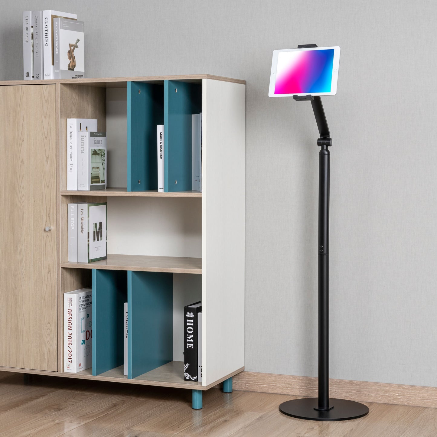 Stabilt gulvstativ til tablet 4,7-12,9" i metal ved siden af hvid-træ reol med bøger og skulptur, lyst trægulv.