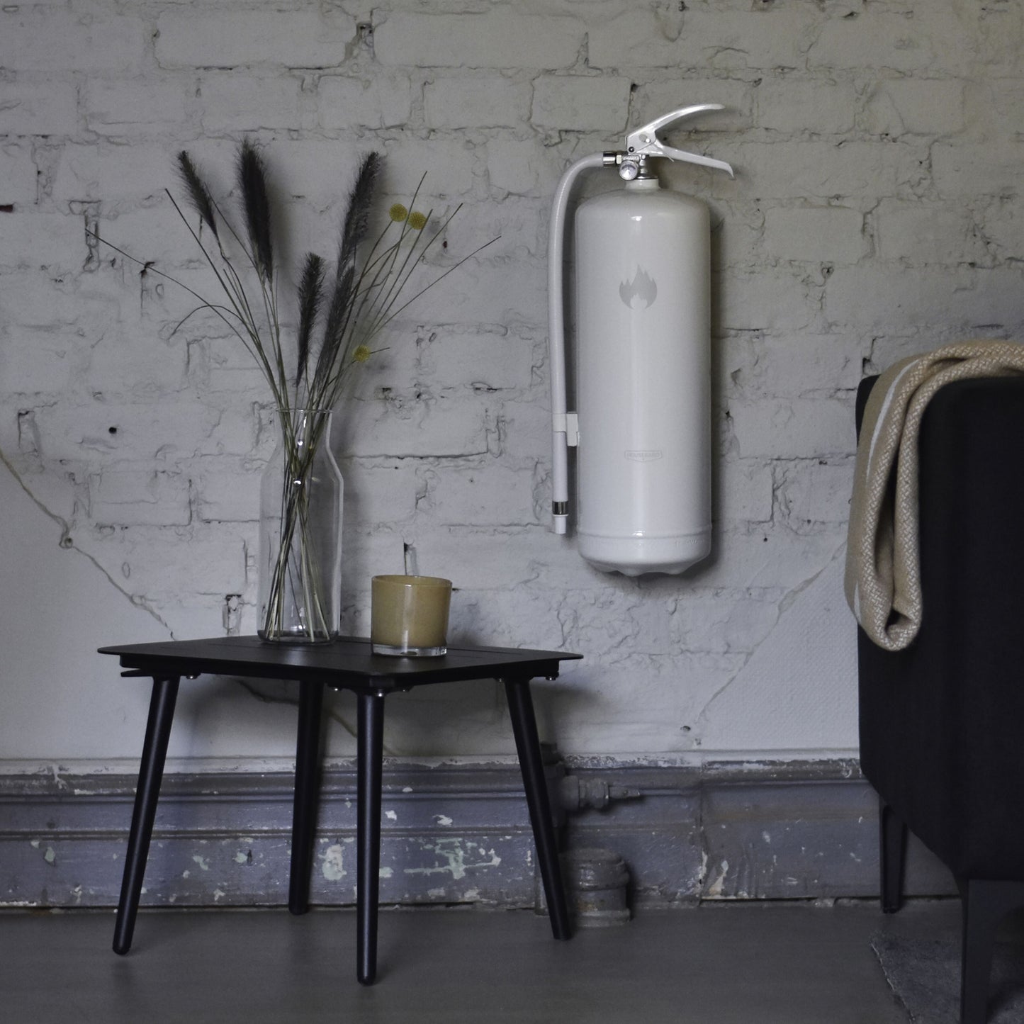 Housegard Brandslukker Design Edition pulver 6 kg hvid vægmonteret over sort bord med lys, vase og delvist synlig mørk sofa.