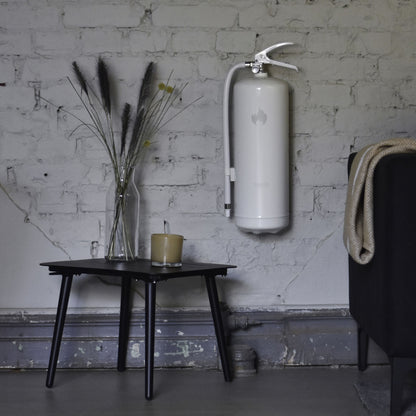 Housegard Brandslukker Design Edition pulver 6 kg hvid vægmonteret over sort bord med lys, vase og delvist synlig mørk sofa.