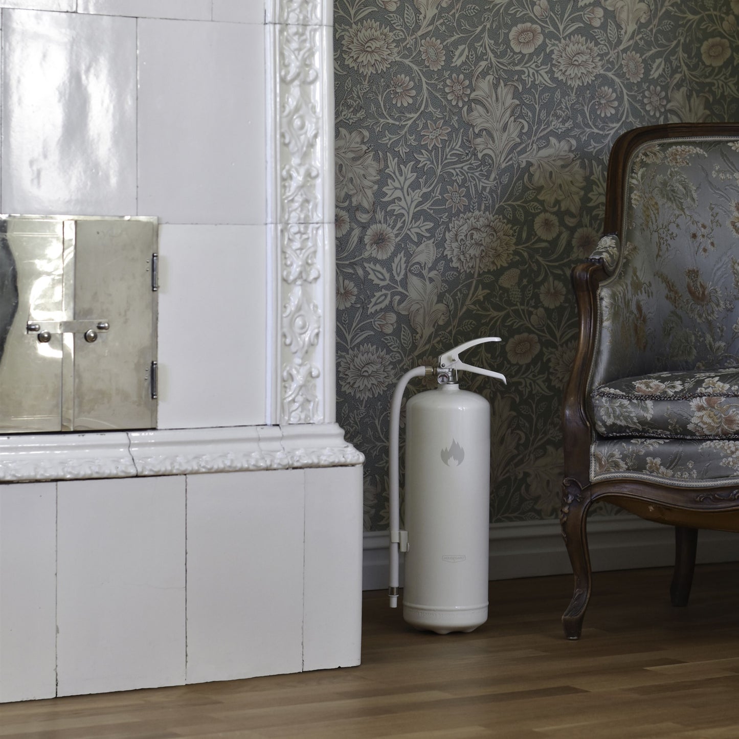 Housegard Brandslukker Design Edition pulver 6 kg hvid ved kakkelpejs, mønstret antik lænestol og blomstret tapet.