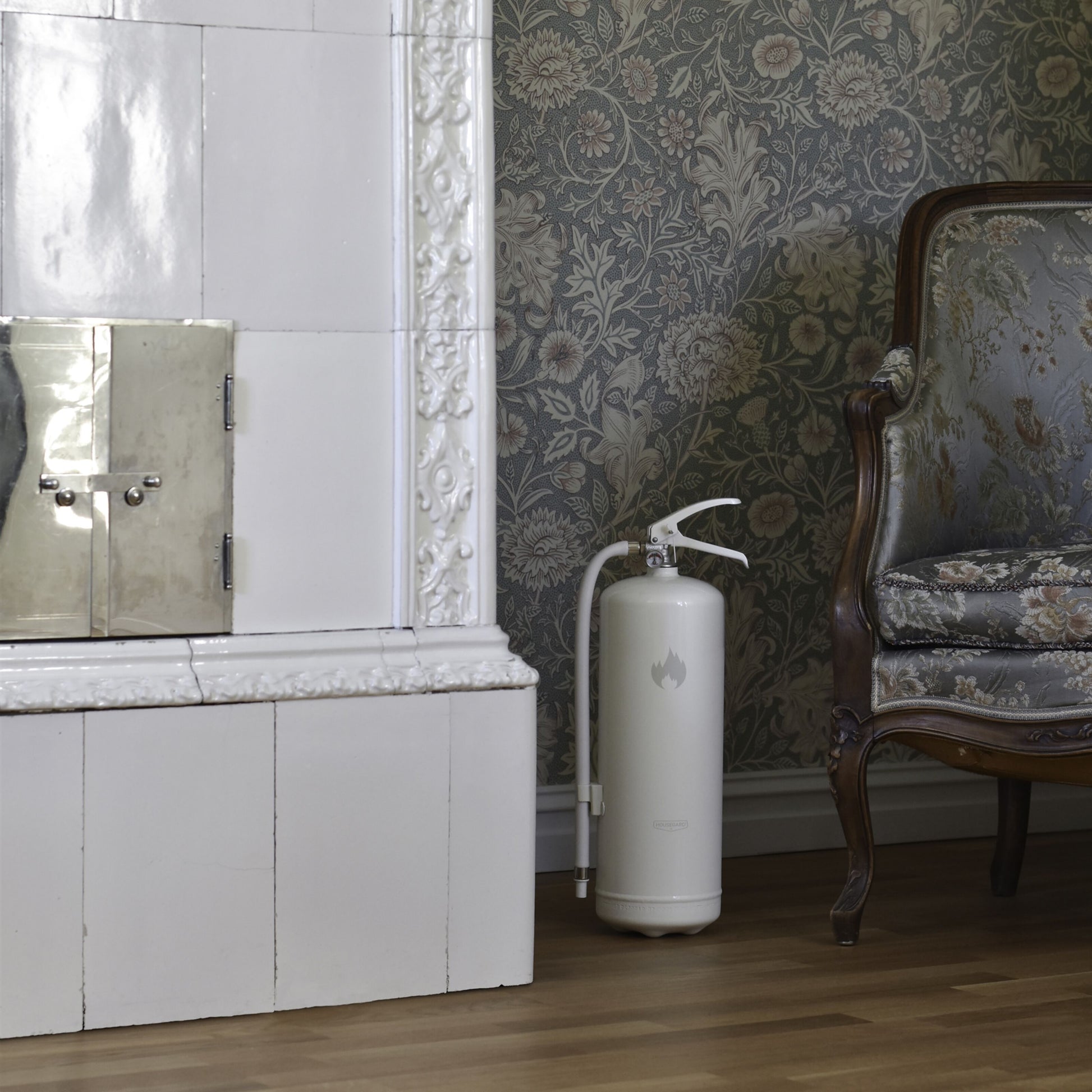 Housegard Brandslukker Design Edition pulver 6 kg hvid ved kakkelpejs, mønstret antik lænestol og blomstret tapet.