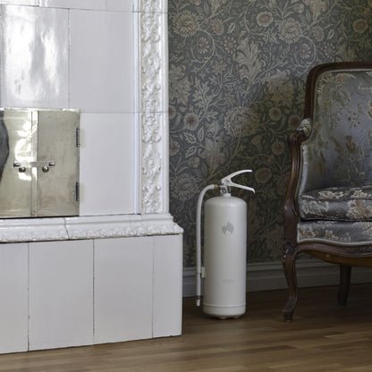 Housegard Brandslukker Design Edition pulver 6 kg hvid ved kakkelpejs, mønstret antik lænestol og blomstret tapet.
