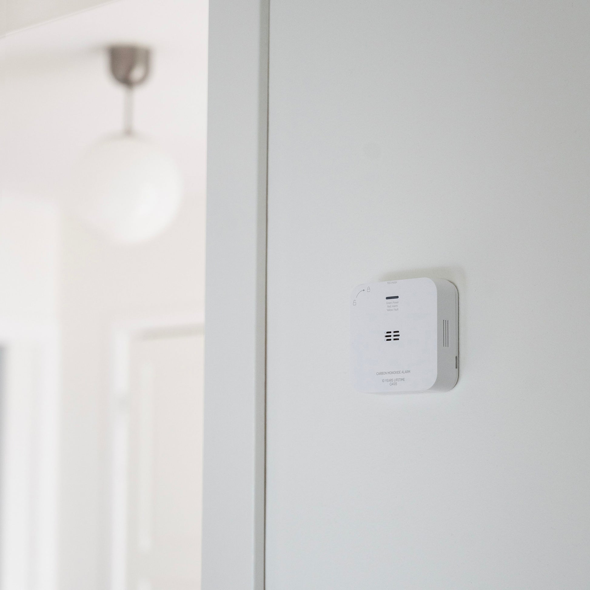 Housegard Luma trådløs kuliltealarm, hvid, monteret på væg i hjem med rund loftlampe og dør synlig i baggrunden.
