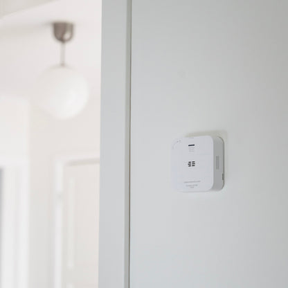 Housegard Luma trådløs kuliltealarm, hvid, monteret på væg i hjem med rund loftlampe og dør synlig i baggrunden.