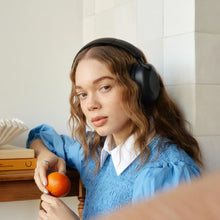 SUDIO K2 trådløse over-ear-hovedtelefoner i sort bæres af en kvinde med bølget hår og en blå kjole, der sidder ved et bord med bøger.