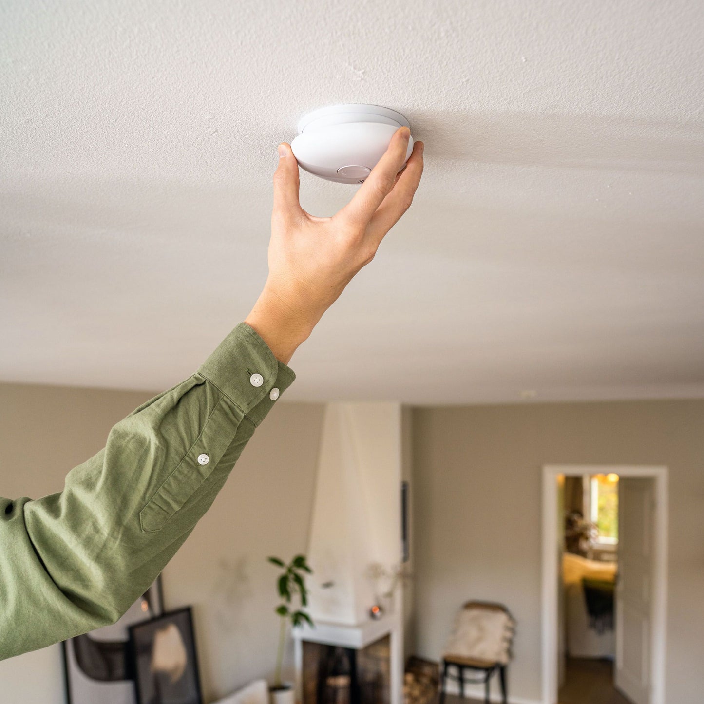 Housegard Pebble 10 optisk røgalarm monteret på hvidt loft, grøn skjorte synlig, lyse vægge, plante i baggrunden.