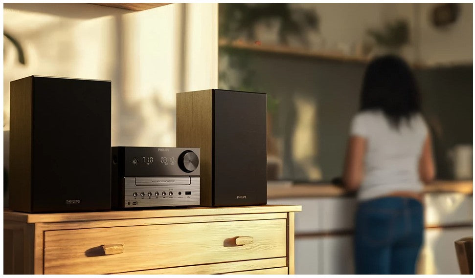 Philips Mikromusiksystem TAM3505 sort med to højttalere på trækommode, sløret person og køkken i baggrunden.
