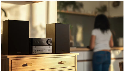 Philips Mikromusiksystem TAM3505 sort med to højttalere på trækommode, sløret person og køkken i baggrunden.