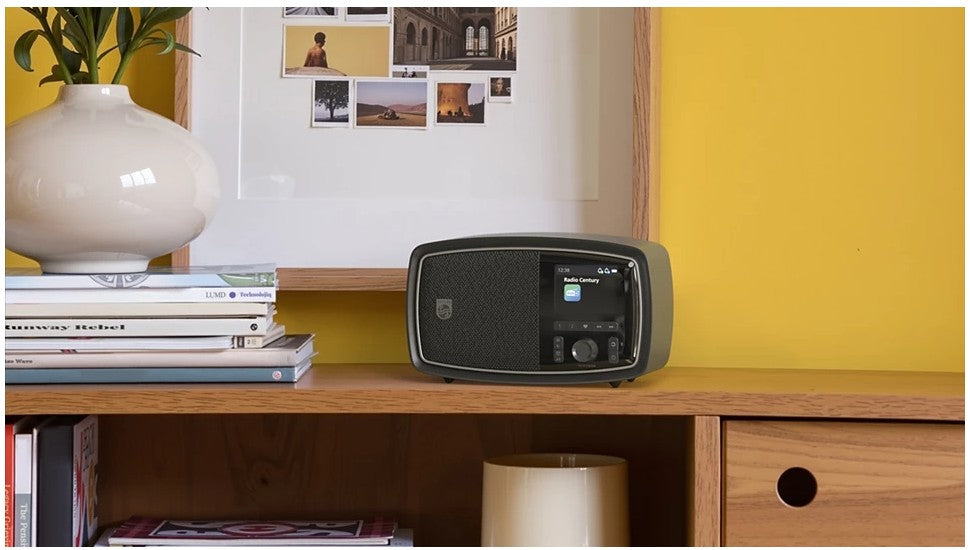 Philips The Janet bærbar retroradio DAB+ Bluetooth på træbord ved bøger og hvid vase nær gul væg med fotocollage.