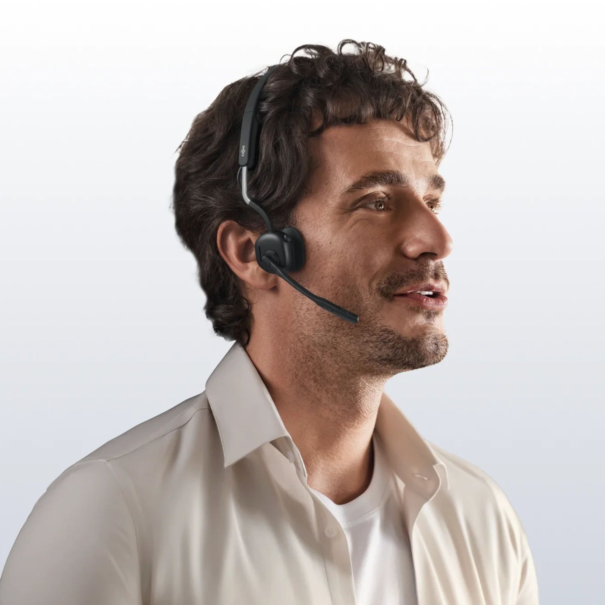 Shokz OpenMeet kontorhovedtelefoner med åbent øre-design, trådløs, mikrofon, bæres af mand med krøllet hår og beige skjorte.
