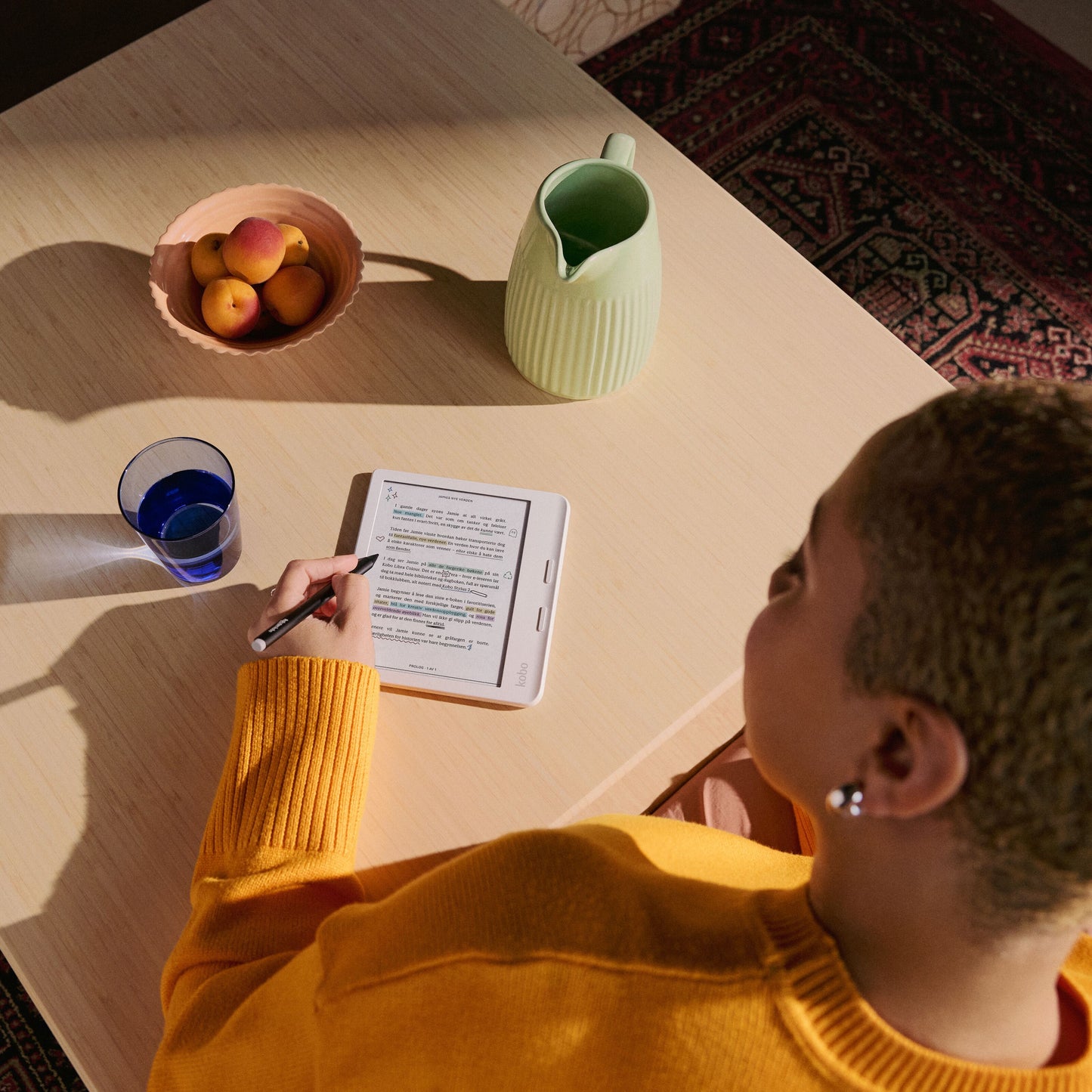 Kobo Libra Colour læseplade 7" Sort med stylus, set fra siden på træbord, delvist synlig frugtskål og gulvtæppe.