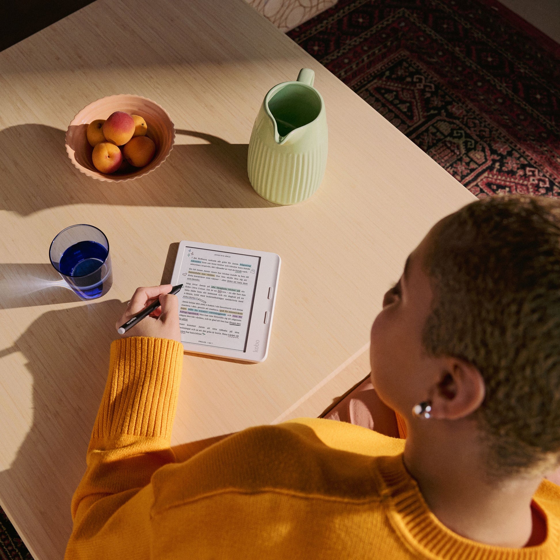 Kobo Libra Colour læseplade 7" Sort med E Ink Kaleido 3, bruges med stylus ved lys trætabel, frugt og glas synlige.