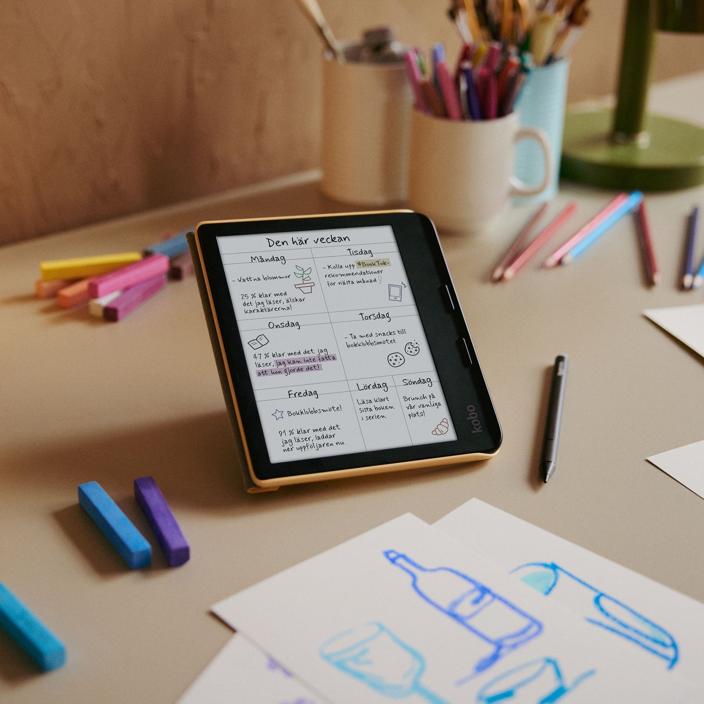 Kobo Libra Colour læseplade 7" Sort med håndskrevet ugemenu, farvet pen og markør, synlig kop og grøn lampe.