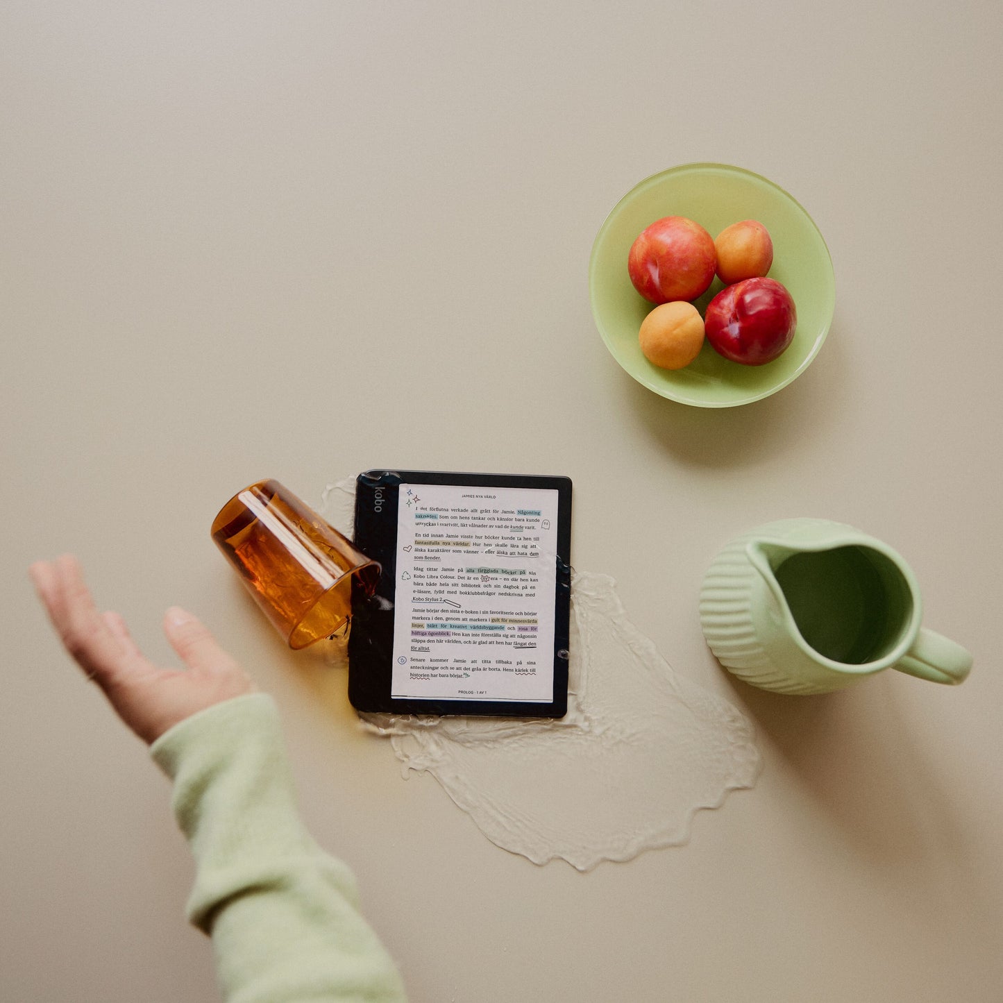 Kobo Libra Colour læseplade 7" Sort på bord, vand spildt fra væltet ravfarvet glas, grøn kande og frugtskål ved siden.
